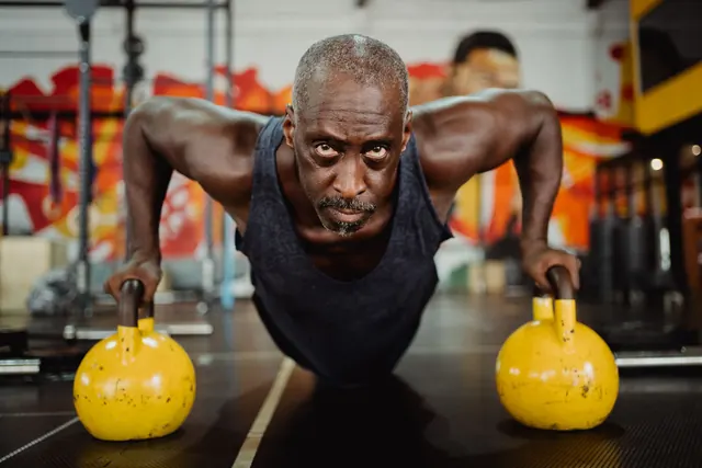 A gray-haired man holding a push-up position on two kettlebells in a gym