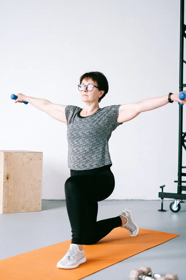 A woman in glasses holding a kneeling lunge with light dumbbells, arms out to the sides