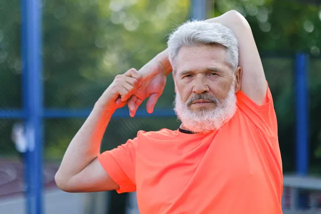 A gray-haired, bearded man stretching his arm overhead outdoors