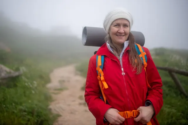 A smiling woman with a backpack and knit hat on a foggy hiking trail